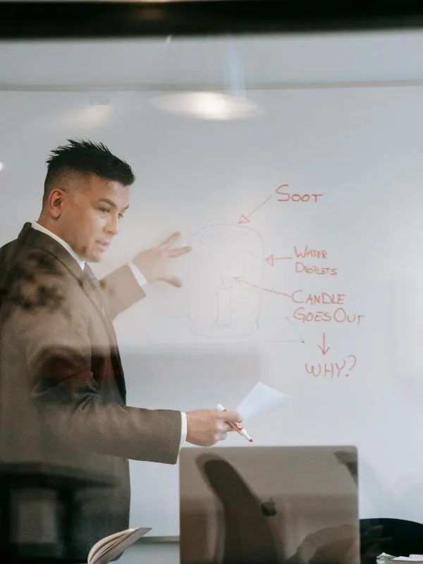 A cheerful middle-aged British man teaching English in front of a camera with whiteboard notes.