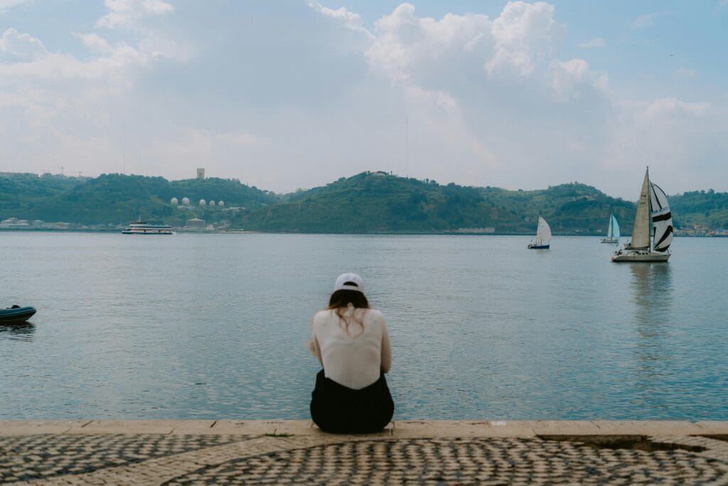 A solo traveler sitting on a balcony in Lisbon with pastel rooftops and the Tagus River in view.